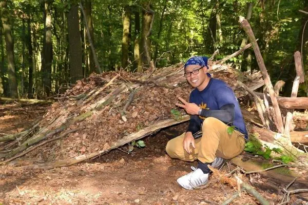 Person zeigt auf selbstgebaute Unterkunft aus Ästen und Blättern
