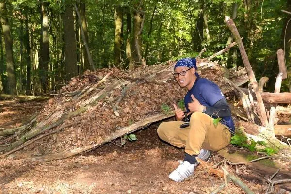 Person zeigt Daumen hoch neben selbstgebautem Unterschlupf aus Ästen