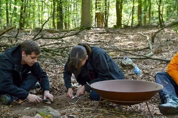 Personen sammeln Materialien neben einer Feuerstelle im Wald beim Survival Training