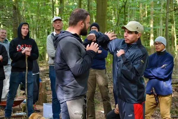 Zwei Personen demonstrieren eine Selbstverteidigungstechnik im Wald, umgeben von Zuschauern