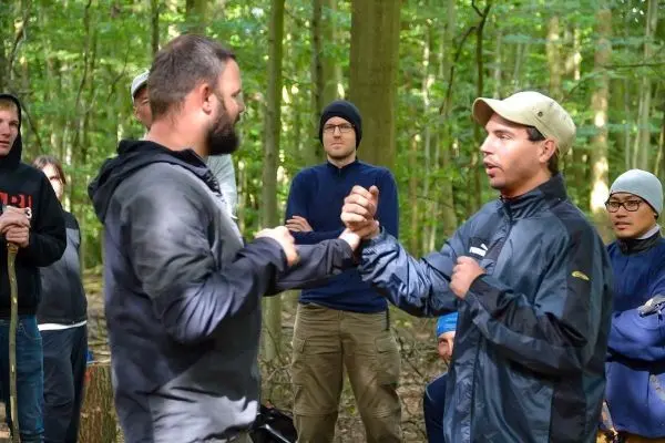 Zwei Personen demonstrieren eine Technik im Wald, Zuschauer beobachten aufmerksam