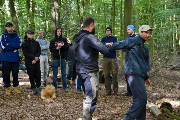 Zwei Personen demonstrieren eine Technik im Wald, Zuschauer beobachten aufmerksam
