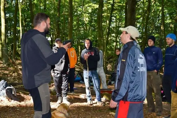 Zwei Personen diskutieren im Wald, während eine Gruppe von Teilnehmern zuschaut