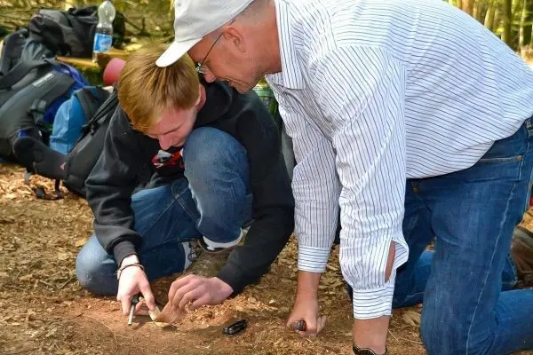 Zwei Personen knien am Boden und bearbeiten Holz mit einem Messer