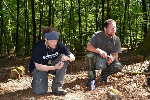 Zwei Personen knien im Wald und bearbeiten Holz mit einem Messer