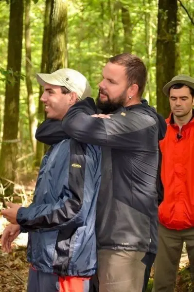 Zwei Personen stehen im Wald, eine Person umarmt die andere von hinten