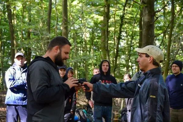 Zwei Personen übergeben sich ein Objekt im Wald, mehrere Personen beobachten
