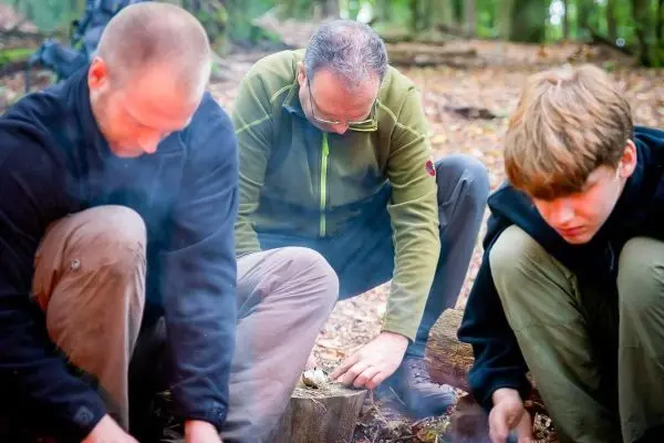 Drei Personen knien am Boden und bereiten Zunder für ein kleines Bodenfeuer vor