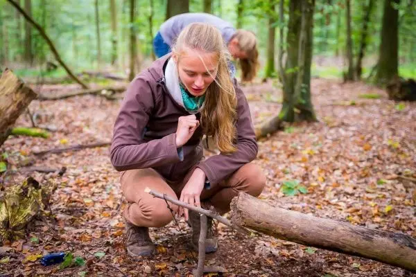 Frau kniet im Wald und bearbeitet einen Stock mit einem Messer