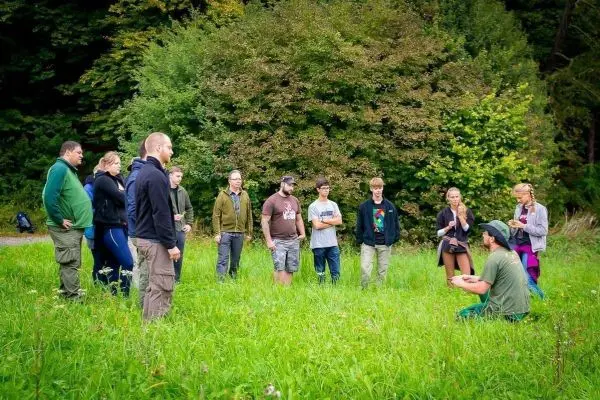 Gruppe steht im Gras und hört einem sitzenden Referenten zu