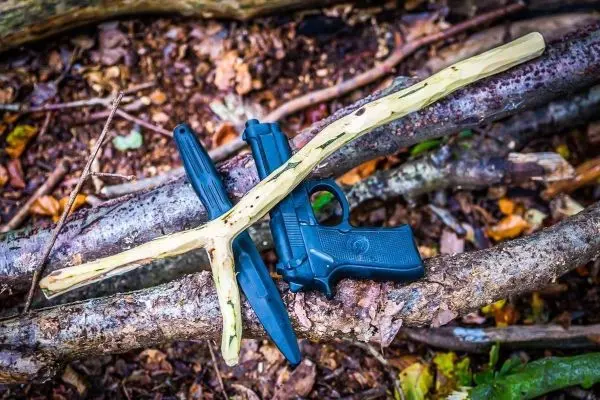 Holzstock und Messer liegen auf Ästen, daneben eine schwarze Pistole