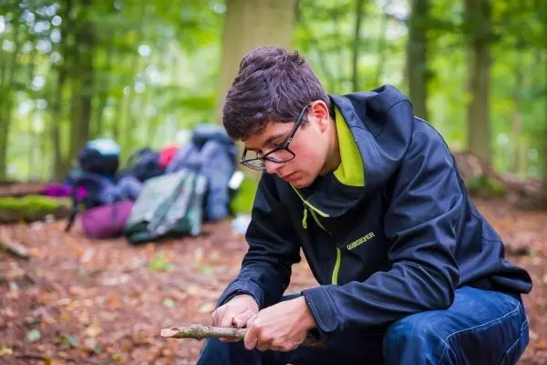 Junge schnitzt mit einem Messer an einem Holzstück im Wald