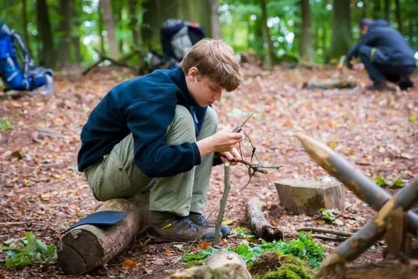 Junge sitzt auf einem Baumstamm und bearbeitet Äste mit einem Messer