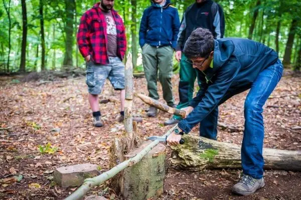 Junger Mann bearbeitet Holzstück mit einem Messer, während andere zuschauen