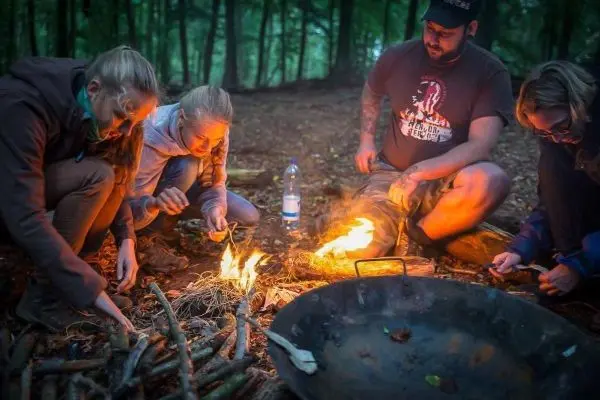 Kleine Flamme brennt zwischen Holzstücken, während mehrere Personen Zunder vorbereiten