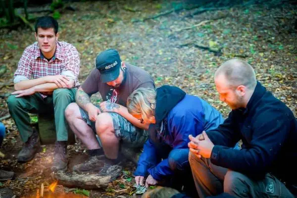 Kreis von Personen sitzt um ein kleines Feuer im Wald, einige halten Materialien in den Händen