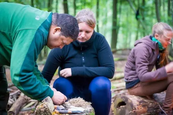 Mann bereitet Zunder auf einem Baumstamm vor, während Frauen im Hintergrund sitzen