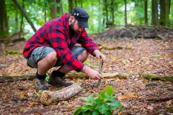 Mann kniet im Wald und bearbeitet einen Stock mit einem Messer