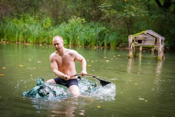 Mann paddelt mit einer improvisierten Bootskonstruktion über einen ruhigen Teich