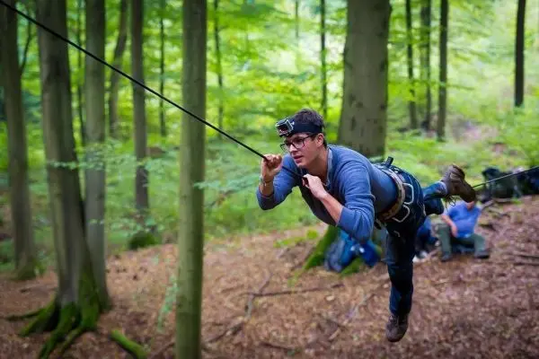 Mann überquert selbstgebaute Seilbrücke im Wald, gesichert mit Gurt und Seil