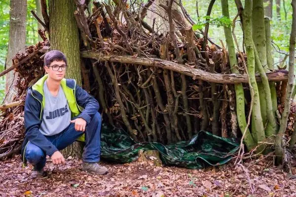 Person sitzt neben einer selbstgebauten Hütte aus Ästen im Wald