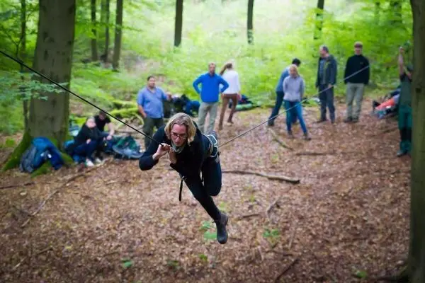 Person überquert selbstgebaute Seilbrücke im Wald, Zuschauer im Hintergrund