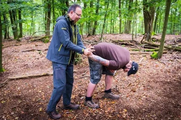 Zwei Personen in einem Wald, eine Person hilft der anderen beim Bücken