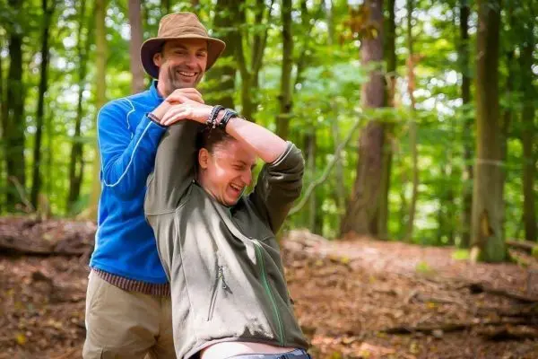 Zwei Personen interagieren spielerisch im Wald, einer hält den anderen am Handgelenk
