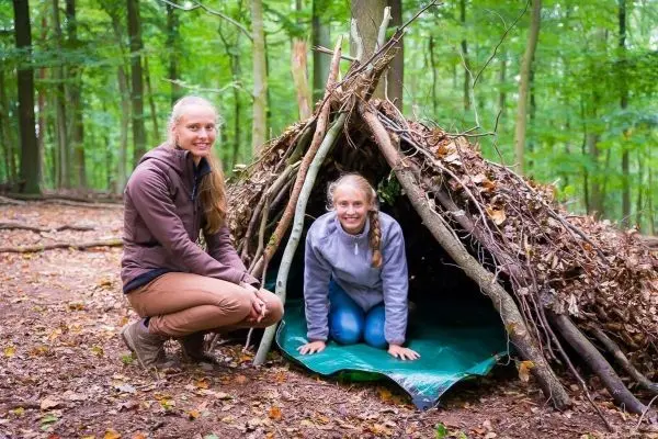 Zwei Personen neben einer selbstgebauten Tarp-Notunterkunft aus Ästen und Blättern