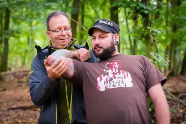 Zwei Personen stehen im Wald und zeigen auf einen Arm mit Tätowierung