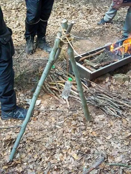 Drei Äste bilden ein Gestell mit einer Wasserflasche, daneben brennt ein Feuer
