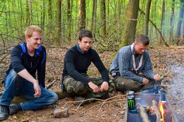 Drei Personen sitzen am Boden und beobachten ein kleines Feuer im Wald