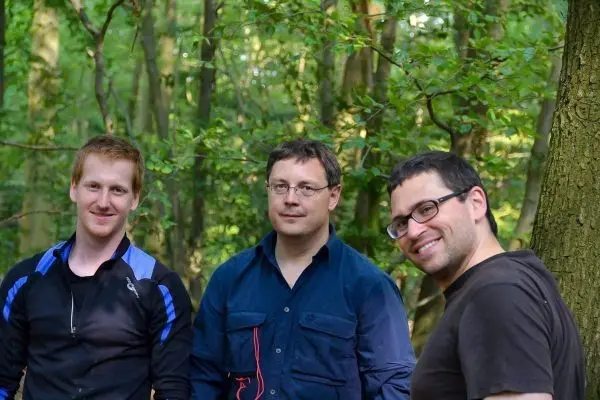 Drei Personen stehen im Wald zwischen Bäumen und lächeln in die Kamera