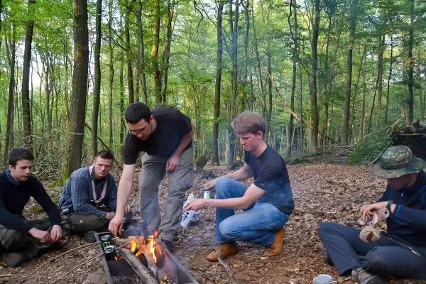 Feuer brennt in einer improvisierten Feuerstelle, während Teilnehmer Zubereitung beobachten