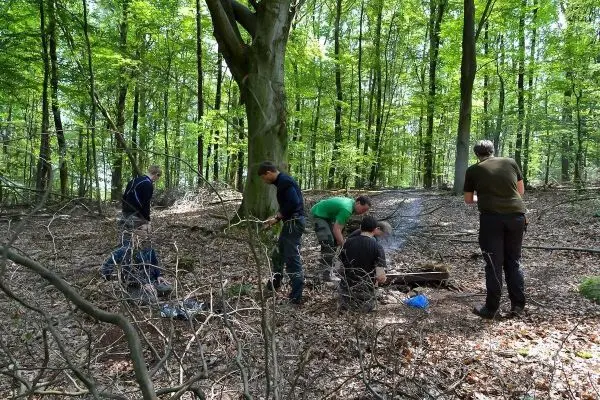 Gruppe arbeitet im Wald an einer Feuerstelle mit Holz und Ausrüstung