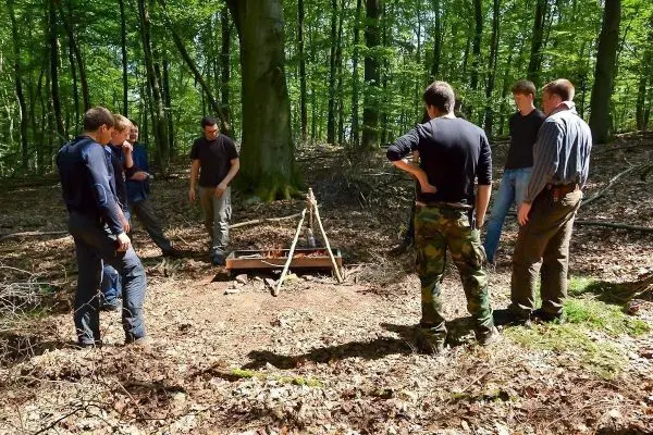 Gruppe betrachtet selbstgebauten Hobo-Kocher unter Bäumen im Wald