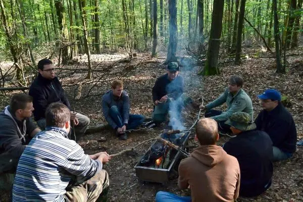 Gruppe sitzt um ein Feuer im Wald und spricht miteinander beim Survival Training