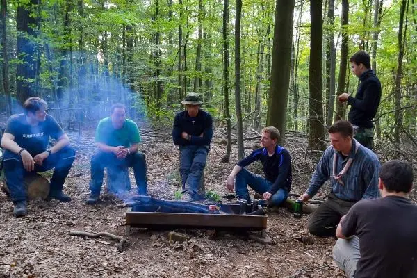 Gruppe sitzt um eine Feuerstelle im Wald und beobachtet das Feuer beim Überlebenstraining