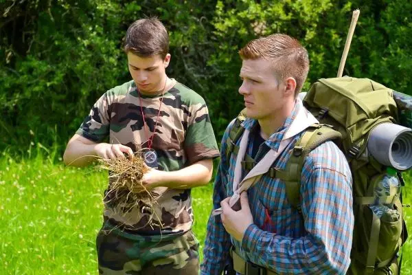 Junger Mann bereitet Zunder aus Pflanzenmaterial vor, während ein anderer mit Rucksack wartet
