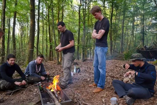 Kleines Bodenfeuer brennt in einer Holzschale, mehrere Personen sitzen und stehen darum herum