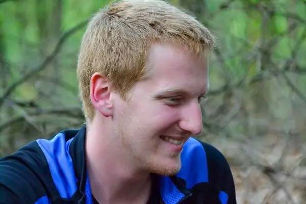 Lächelnder junger Mann sitzt im Wald, umgeben von Ästen und Laub