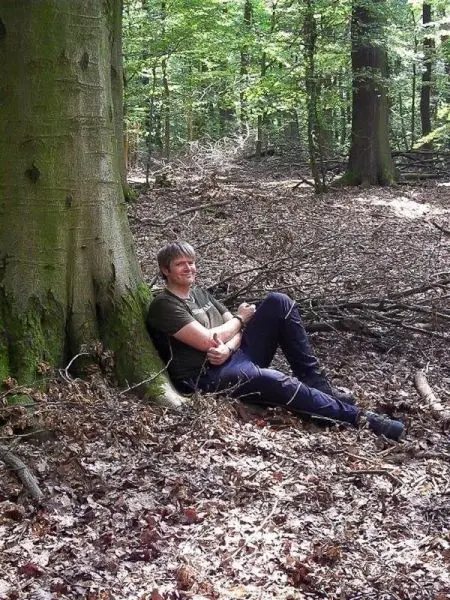 Mann lehnt entspannt an einem Baumstamm im Wald auf Laub