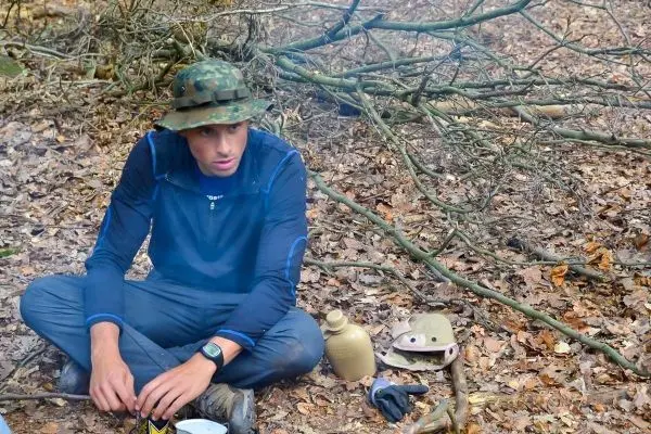 Mann sitzt auf dem Boden im Wald neben einem Kochtopf und einer Wasserflasche