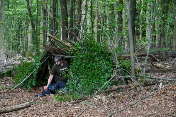 Mann sitzt vor einer selbstgebauten Tarp-Notunterkunft im Wald