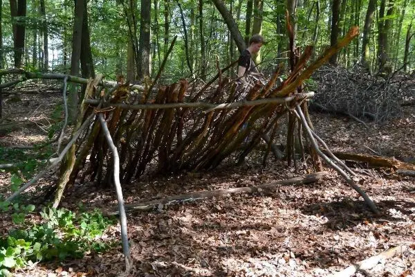 Selbstgebaute Laubhütte aus Ästen im Wald, Person steht daneben