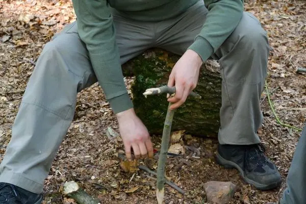 Stock wird mit einem Messer bearbeitet, während auf einem Baumstumpf gesessen wird