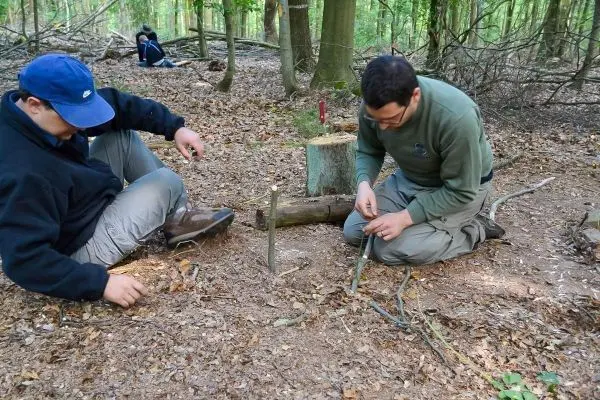 Zwei Personen bearbeiten Holzstücke auf dem Waldboden beim Überlebenstraining