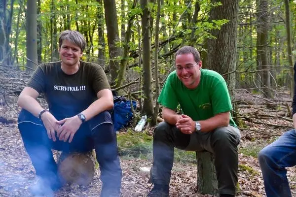 Zwei Personen sitzen auf Baumstümpfen im Wald, Rauch steigt auf