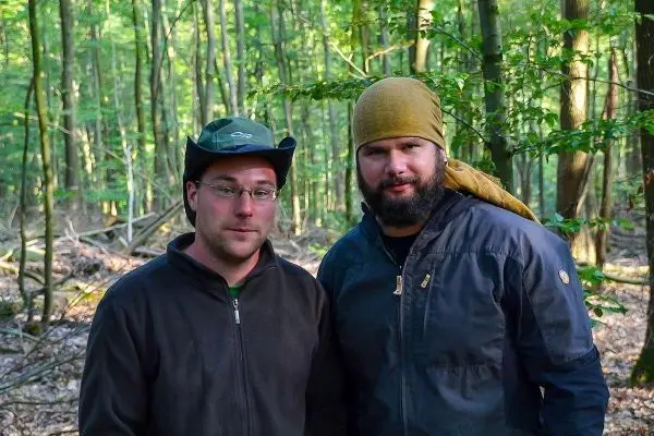 Zwei Personen stehen im Wald zwischen Bäumen und Laub