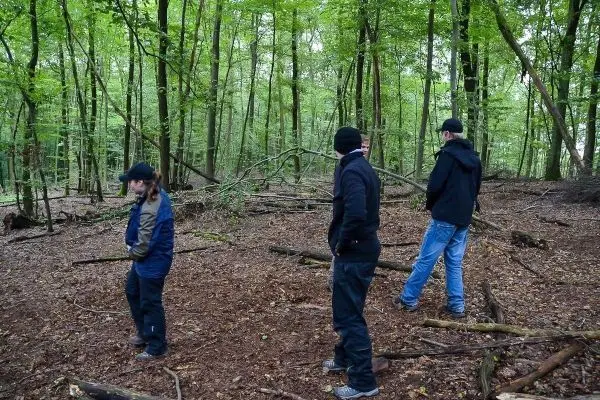 Drei Personen bewegen sich durch einen bewaldeten Bereich mit Laub und Ästen